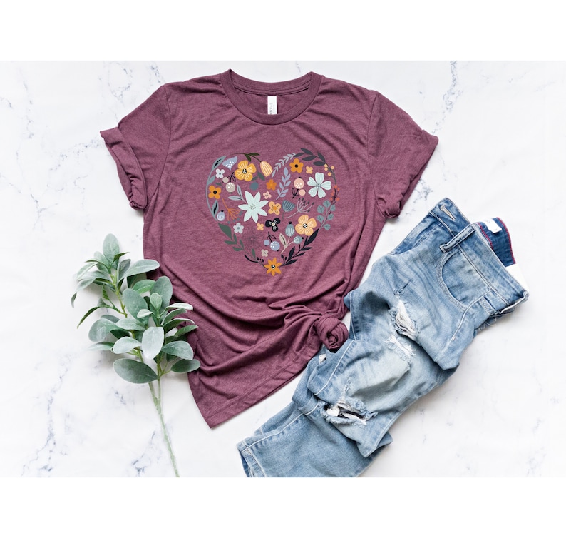 K&ouml;nnte beinhalten: Ein maroonfarbenes T-Shirt mit einem floralen Herz-Design. Das Herz besteht aus verschiedenen Blumen in Blaut&ouml;nen, Gelb und Gr&uuml;n. Das Shirt wird auf einer wei&szlig;en Marmorfl&auml;che mit einem Zweig Gr&uuml;n und einer blauen Jeans gezeigt.