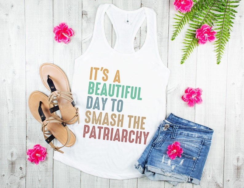 以下が含まれることがあります： カラフルな文字で「It's a beautiful day to smash the patriarchy」と書かれた白いタンクトップ。タンクトップは、白い木製の表面に、サンダル、デニムのショートパンツ、ピンクの花と一緒に置かれています。