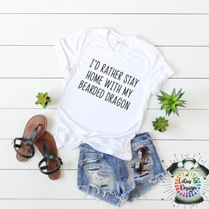 Puede incluir: Camiseta blanca con el texto "I'd rather stay home with my bearded dragon". La camiseta est&aacute; sobre una superficie de madera blanca con un par de sandalias marrones, pantalones cortos de mezclilla azul, gafas de sol y dos peque&ntilde;as plantas verdes.