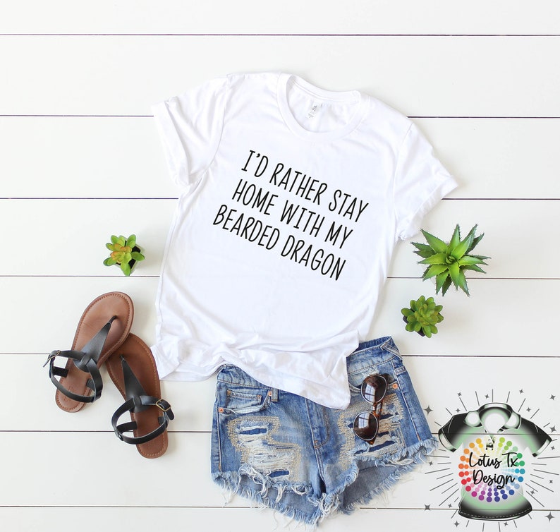 Puede incluir: Camiseta blanca con el texto "I'd rather stay home with my bearded dragon". La camiseta est&aacute; sobre una superficie de madera blanca con un par de sandalias marrones, pantalones cortos de mezclilla azul, gafas de sol y dos peque&ntilde;as plantas verdes.