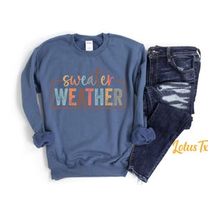 May include: A blue sweatshirt with the text "sweater weather" in colorful letters. The sweatshirt is laid flat on a white surface with a pair of blue jeans next to it.