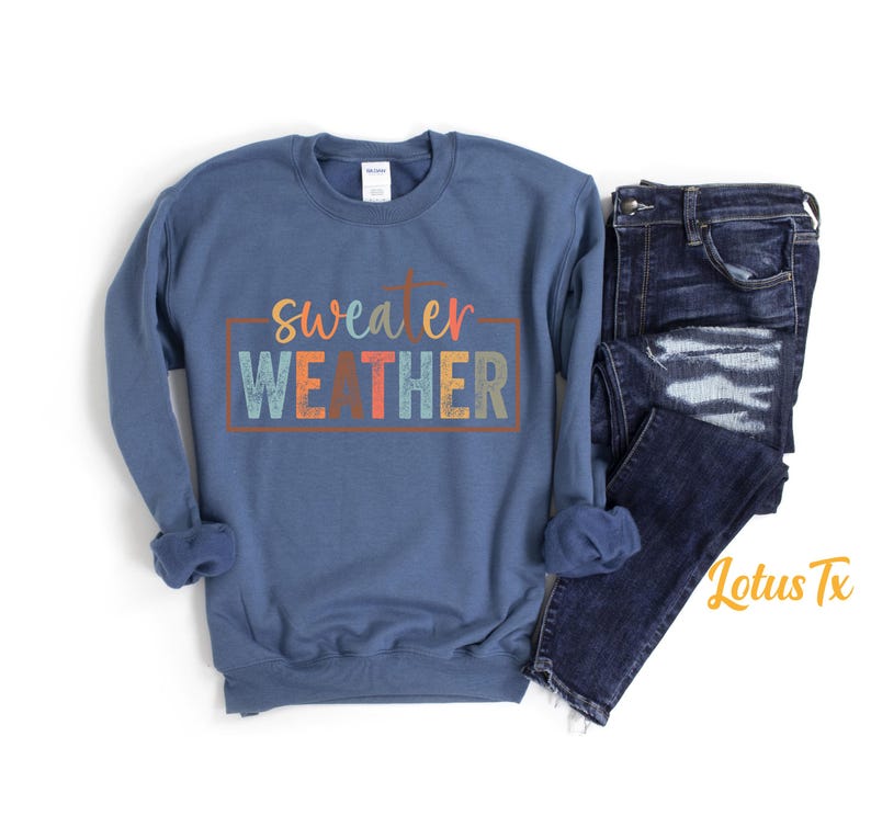 May include: A blue sweatshirt with the text "sweater weather" in colorful letters. The sweatshirt is laid flat on a white surface with a pair of blue jeans next to it.