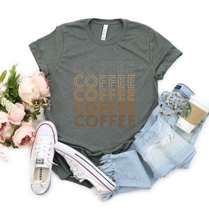 K&ouml;nnte beinhalten: Ein gr&uuml;nes T-Shirt mit dem Wort "COFFEE" f&uuml;nfmal in einer Retro-Schriftart wiederholt. Das Wort "COFFEE" ist in verschiedenen Braunt&ouml;nen gehalten. Das T-Shirt liegt auf einer wei&szlig;en Oberfl&auml;che mit einem Paar wei&szlig;en Sneakers, einer blauen Jeans und einer Kaffeetasse.