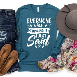 May include: Teal long-sleeve shirt with white text that reads "Everyone was thinking it I just said it". The shirt is paired with blue jeans, brown flats, a floral scarf, and a gray fedora.