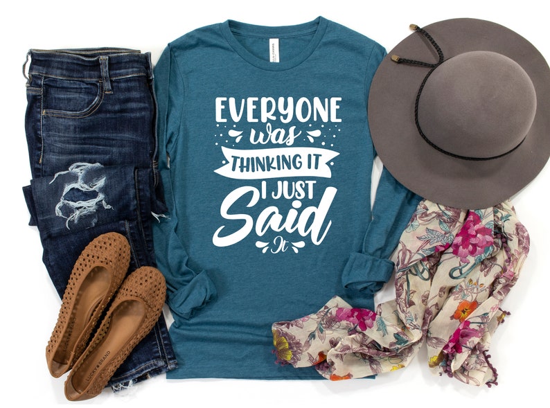 May include: Teal long-sleeve shirt with white text that reads "Everyone was thinking it I just said it". The shirt is paired with blue jeans, brown flats, a floral scarf, and a gray fedora.