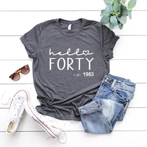 May include: A dark grey t-shirt with the text "hello forty est. 1983" in white. The shirt is laid out on a white wooden surface with a pair of sunglasses, a pair of jeans, and a pair of white sneakers.