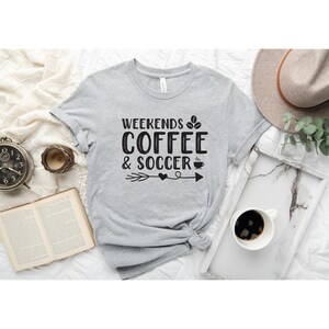 Op de afbeelding: Grijs T-shirt met de tekst "Weekends Coffee & Soccer" in zwarte letters met een afbeelding van een koffiekopje en een voetbal.