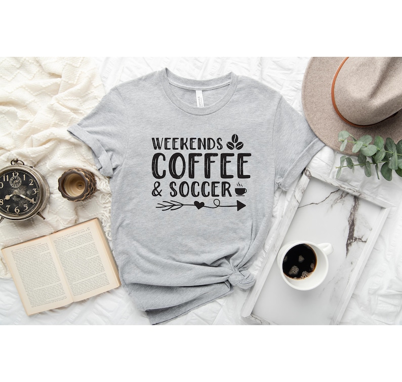 Op de afbeelding: Grijs T-shirt met de tekst "Weekends Coffee & Soccer" in zwarte letters met een afbeelding van een koffiekopje en een voetbal.