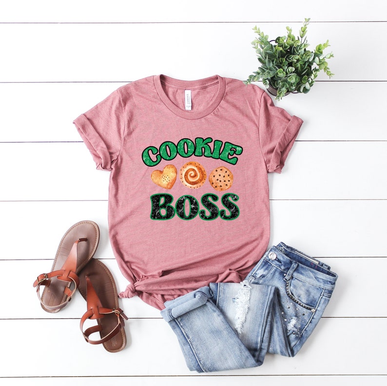 K&ouml;nnte beinhalten: Ein pinkfarbenes T-Shirt mit dem Text "Cookie Boss" in gr&uuml;nen und schwarzen Buchstaben. Drei Kekse sind &uuml;ber dem Text abgebildet.