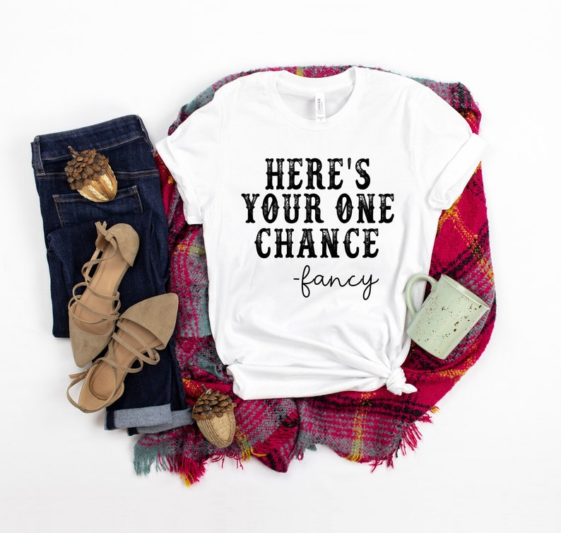 May include: White t-shirt with the text "Here's your one chance -fancy" in black lettering. The shirt is laid out on a red and white plaid blanket with a pair of brown sandals, blue jeans, and a small green mug.