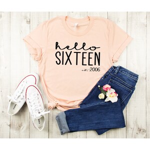 May include: A peach-colored t-shirt with the text "hello sixteen est. 2006" in black. The shirt is paired with blue jeans and white sneakers.
