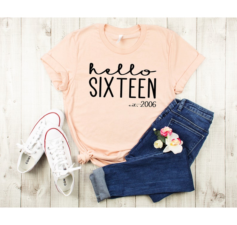 May include: A peach-colored t-shirt with the text "hello sixteen est. 2006" in black. The shirt is paired with blue jeans and white sneakers.