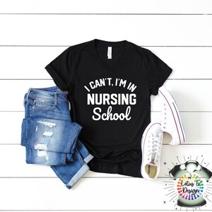 May include: Black v-neck t-shirt with the text "I can't, I'm in nursing school" in white lettering. The shirt is displayed on a white wooden background with a pair of blue jeans and white sneakers.
