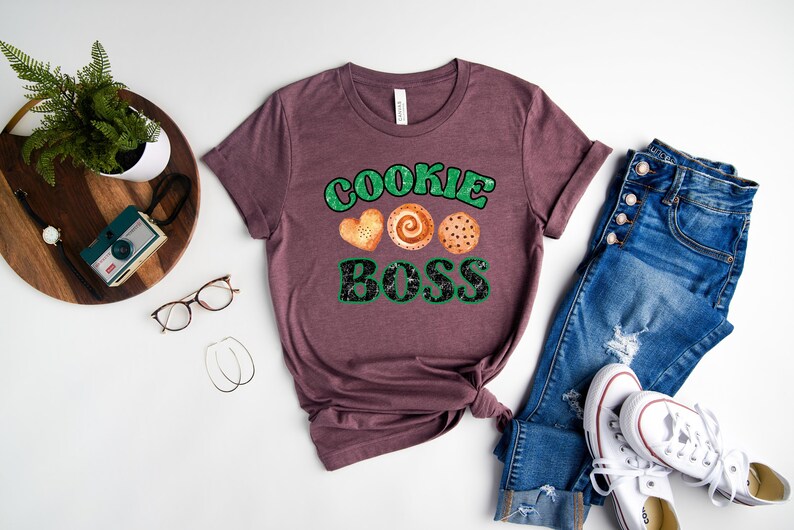 K&ouml;nnte beinhalten: Ein mauvefarbenes T-Shirt mit dem Text "Cookie Boss" in gr&uuml;ner und schwarzer Schrift. Drei Kekse sind &uuml;ber dem Text abgebildet.