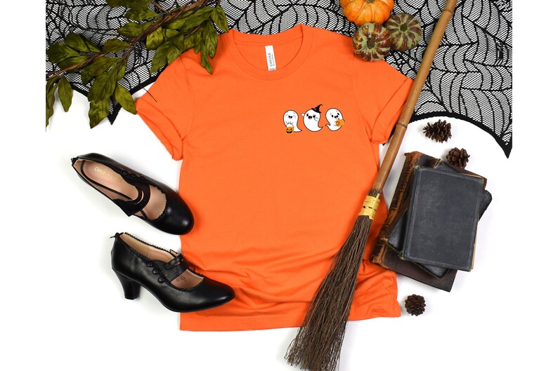 K&ouml;nnte beinhalten: Orangefarbenes T-Shirt mit einem Motiv von drei Geistern, die K&uuml;rbisse halten. Die Geister tragen H&uuml;te und haben unterschiedliche Gesichtsausdr&uuml;cke.