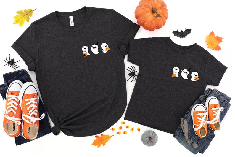 K&ouml;nnte beinhalten: Zwei dunkelgraue T-Shirts mit einem niedlichen Geistermotiv. Das gr&ouml;&szlig;ere Shirt hat drei Geister, die S&uuml;&szlig;mais halten. Das kleinere Shirt hat drei Geister, die S&uuml;&szlig;mais halten. Die Shirts sind von Halloween-Dekorationen umgeben, darunter S&uuml;&szlig;mais, ein K&uuml;rbis und eine Fledermaus.
