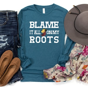 May include: Teal long-sleeve shirt with white text that reads "Blame it all on my roots" with a graphic of a cowboy boot and hat. The shirt is displayed with jeans, a floral scarf, and a brown hat.