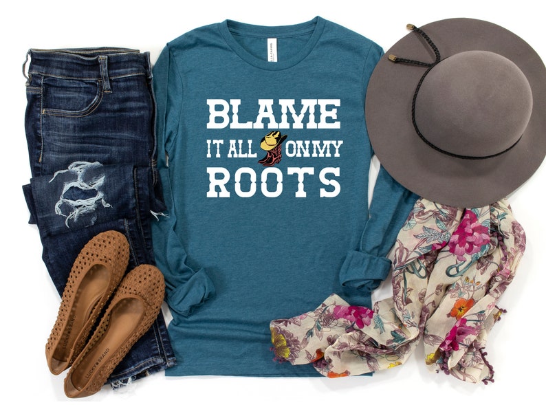 May include: Teal long-sleeve shirt with white text that reads "Blame it all on my roots" with a graphic of a cowboy boot and hat. The shirt is displayed with jeans, a floral scarf, and a brown hat.