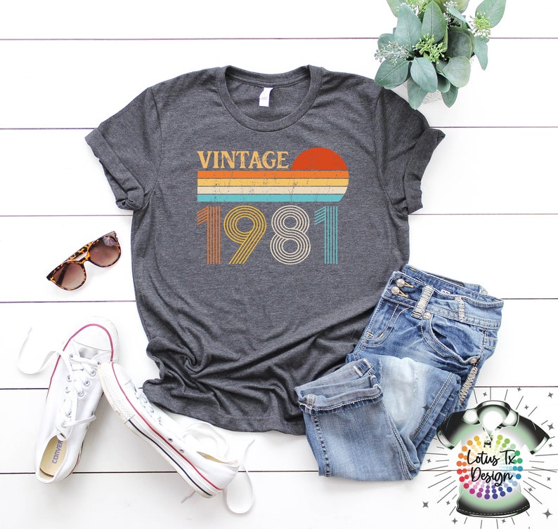 May include: A dark gray t-shirt with a vintage 1981 design featuring a sunset and rainbow stripes. The shirt is laid flat on a white wooden surface with a pair of white sneakers, blue jeans, and sunglasses.