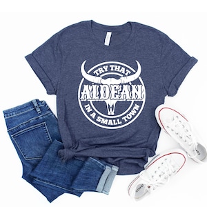 May include: A navy blue t-shirt with a white graphic of a bull skull with the text "Try That In A Small Town" and "ALDEAN" in a circle around the skull.