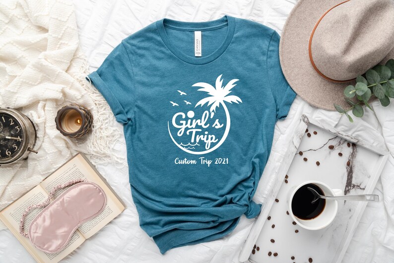 Puede incluir: Camiseta de color turquesa con un gr&aacute;fico blanco que dice "Girl's Trip" con una palmera y p&aacute;jaros. El texto "Custom Trip 2021" est&aacute; debajo del texto principal. La camiseta est&aacute; sobre una superficie blanca con una taza de caf&eacute;, un sombrero y otros accesorios.