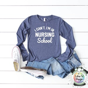 May include: A navy blue long-sleeved shirt with the text "I can't, I'm in nursing school" in white lettering. The shirt is laid out on a white wooden surface with a pair of white sneakers and blue jeans.