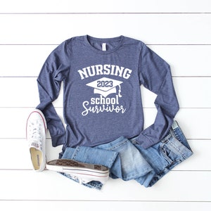 May include: A navy blue long-sleeve shirt with the text "NURSING 2023 school Survivor" in white. The shirt is paired with denim shorts and white sneakers. The items are arranged on a white wooden surface.