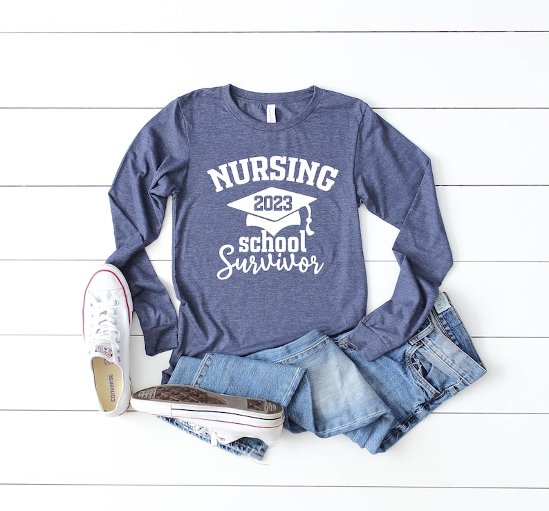 May include: A navy blue long-sleeve shirt with the text "NURSING 2023 school Survivor" in white. The shirt is paired with denim shorts and white sneakers. The items are arranged on a white wooden surface.