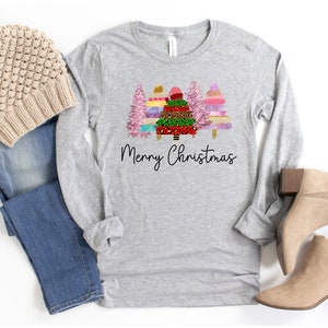 Puede incluir: Una camiseta gris de manga larga con un dise&ntilde;o de tres &aacute;rboles de Navidad coloridos y el texto "Merry Christmas". La camiseta se combina con jeans azules, una boina beige y botines marrones.