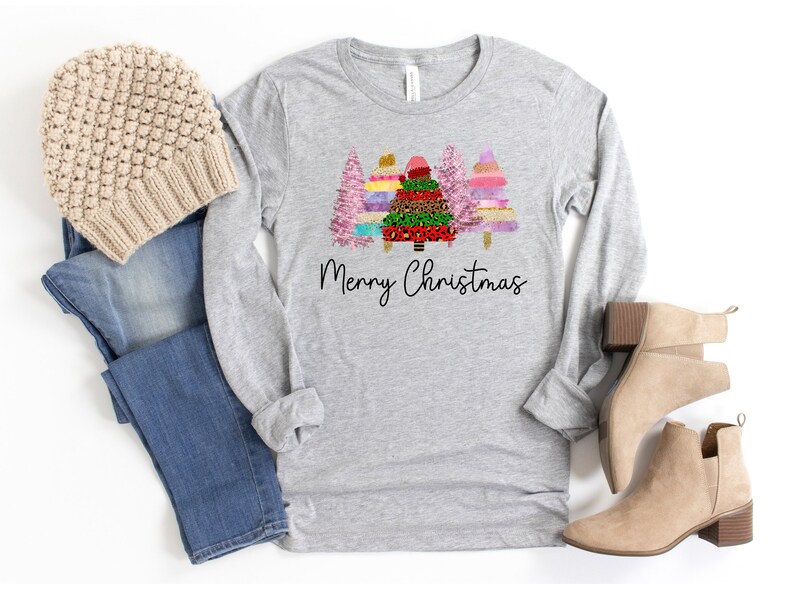 Puede incluir: Una camiseta gris de manga larga con un dise&ntilde;o de tres &aacute;rboles de Navidad coloridos y el texto "Merry Christmas". La camiseta se combina con jeans azules, una boina beige y botines marrones.