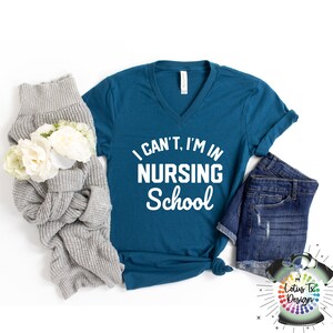 May include: Teal blue v-neck t-shirt with the text "I can't, I'm in Nursing School" in white lettering.