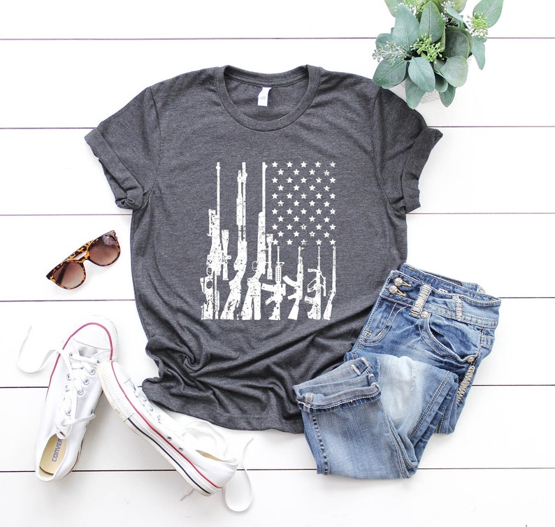 May include: A dark heather grey t-shirt with a white graphic of an American flag with guns printed on it.
