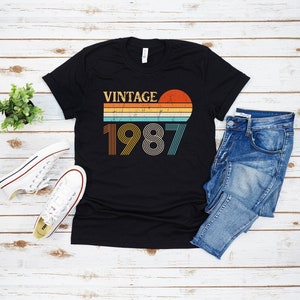 Peut inclure: T-shirt noir avec un design graphique vintage 1987 repr&eacute;sentant un coucher de soleil et des rayures arc-en-ciel. Le texte "Vintage" est au-dessus de l'ann&eacute;e.