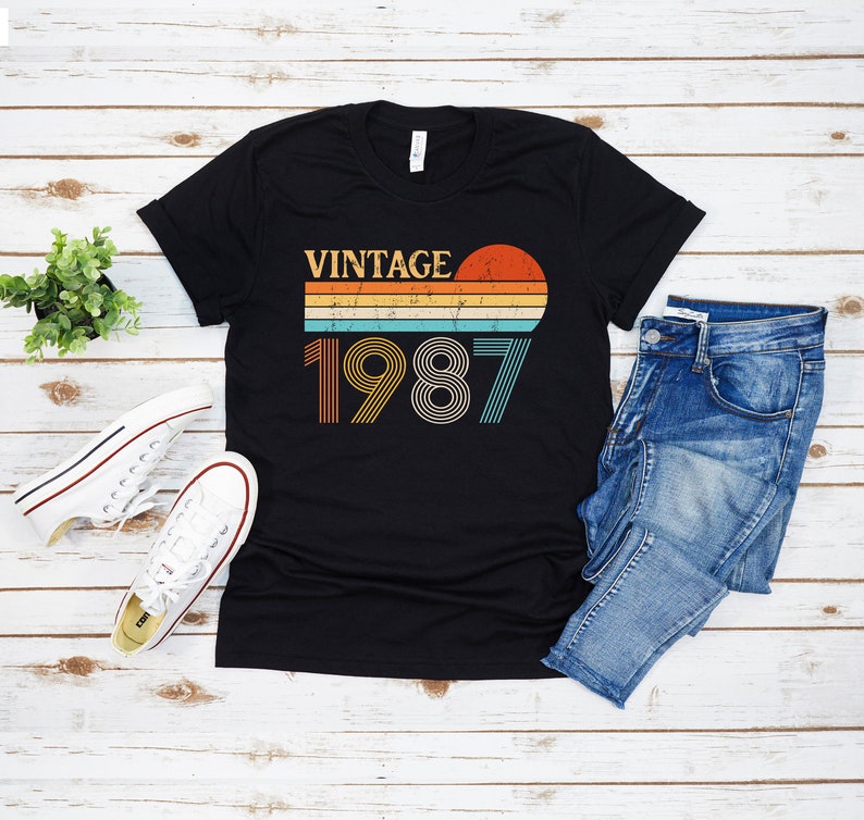 Peut inclure: T-shirt noir avec un design graphique vintage 1987 repr&eacute;sentant un coucher de soleil et des rayures arc-en-ciel. Le texte "Vintage" est au-dessus de l'ann&eacute;e.