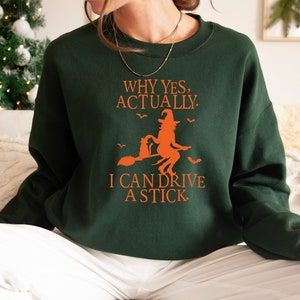 May include: A green sweatshirt with an orange graphic of a witch riding a broomstick. The text on the sweatshirt reads "Why yes, actually. I can drive a stick."