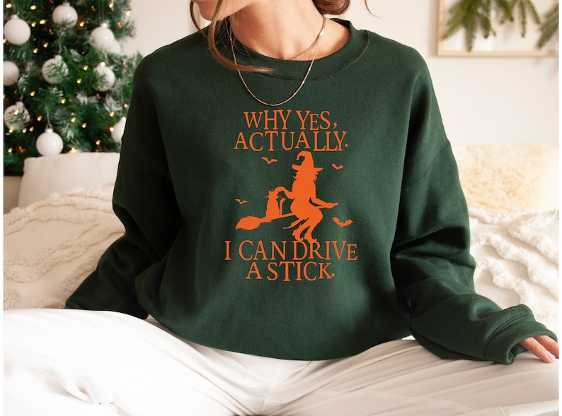 May include: A green sweatshirt with an orange graphic of a witch riding a broomstick. The text on the sweatshirt reads "Why yes, actually. I can drive a stick."