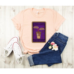 Puede incluir: Una camiseta de color melocot&oacute;n con un gr&aacute;fico de una carta de tarot que presenta una bebida de caf&eacute; helado. La carta est&aacute; etiquetada como "The Iced Coffee".