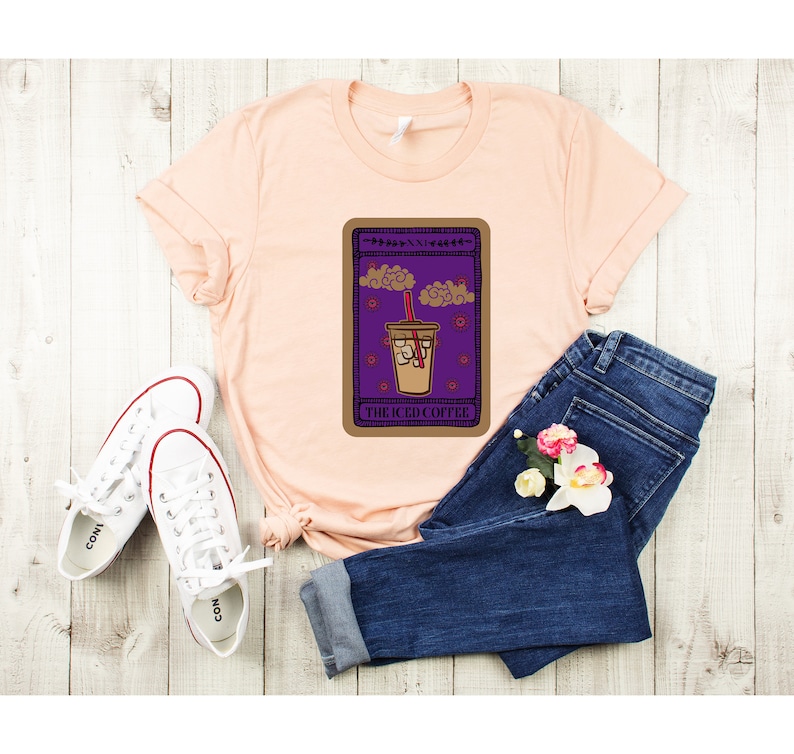 Puede incluir: Una camiseta de color melocot&oacute;n con un gr&aacute;fico de una carta de tarot que presenta una bebida de caf&eacute; helado. La carta est&aacute; etiquetada como "The Iced Coffee".
