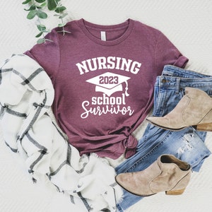 May include: A maroon t-shirt with the text "NURSING 2023 school Survivor" in white, paired with blue jeans, tan ankle boots, and a white and black plaid scarf. The shirt features a graduation cap graphic.