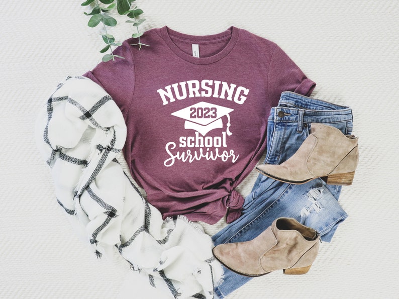 May include: A maroon t-shirt with the text "NURSING 2023 school Survivor" in white, paired with blue jeans, tan ankle boots, and a white and black plaid scarf. The shirt features a graduation cap graphic.