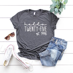 May include: A dark gray t-shirt with the text "hello twenty-five est. 1996" in white. The shirt is laid out on a white wooden surface with a pair of sunglasses, a pair of white sneakers, and a pair of blue jeans.