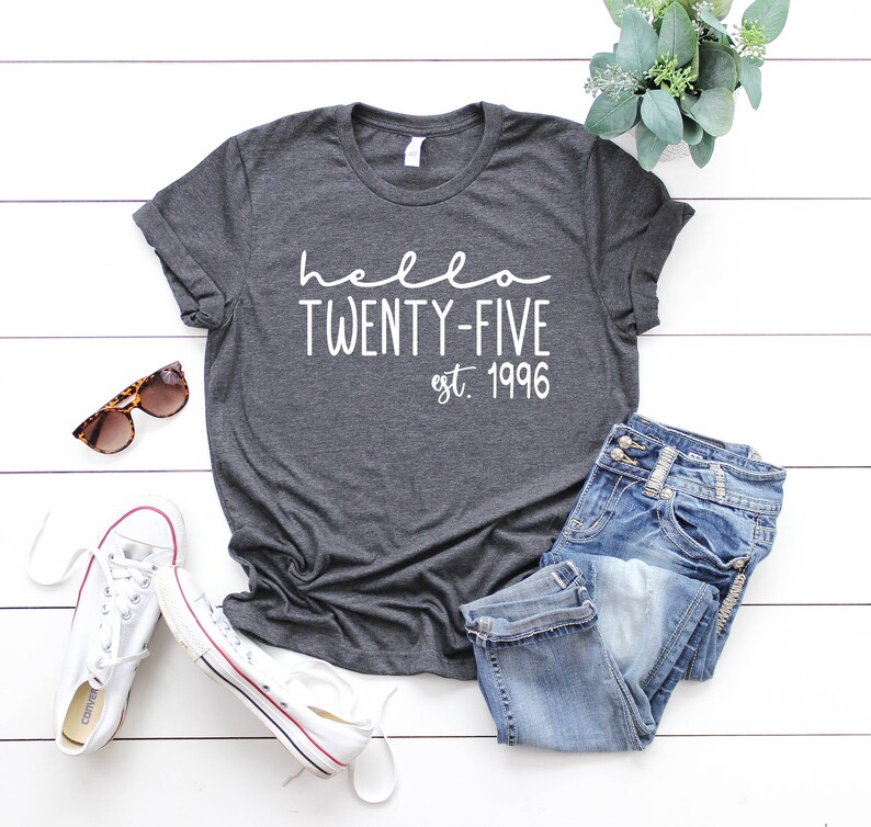 May include: A dark gray t-shirt with the text "hello twenty-five est. 1996" in white. The shirt is laid out on a white wooden surface with a pair of sunglasses, a pair of white sneakers, and a pair of blue jeans.