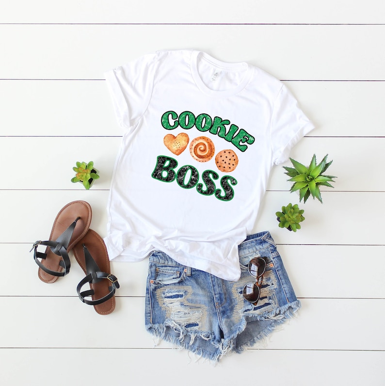 K&ouml;nnte beinhalten: Wei&szlig;es T-Shirt mit dem Text "COOKIE BOSS" in gr&uuml;nen und schwarzen Buchstaben mit einem Herz und zwei Keksdesigns.