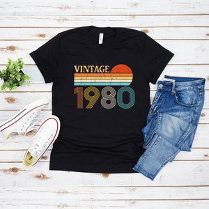 May include: Black t-shirt with a vintage 1980 design featuring a sunset and rainbow stripes. The text "Vintage" is above the year.
