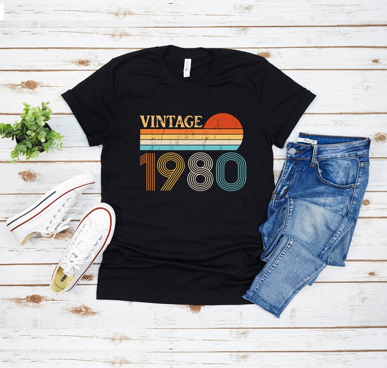 May include: Black t-shirt with a vintage 1980 design featuring a sunset and rainbow stripes. The text "Vintage" is above the year.