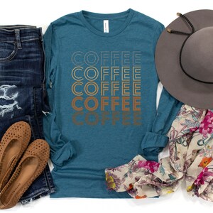 K&ouml;nnte beinhalten: Ein blaues Langarmshirt mit dem Wort "COFFEE", das f&uuml;nfmal in einer Retro-Schrift wiederholt wird. Das Shirt wird mit einer blauen Jeans, braunen Ballerinas, einem floralen Schal und einem braunen Fedora kombiniert.