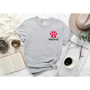 May include: Gray t-shirt with a red paw print with a white plus sign in the center. The text "MEDIC" is printed below the paw print.