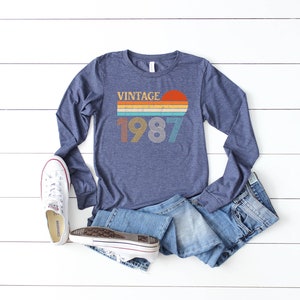 Peut inclure: Un t-shirt &agrave; manches longues bleu marine avec un motif vintage repr&eacute;sentant l'ann&eacute;e 1987 dans une police r&eacute;tro color&eacute;e. Le motif comprend un coucher de soleil avec des rayures orange, jaunes et bleues.