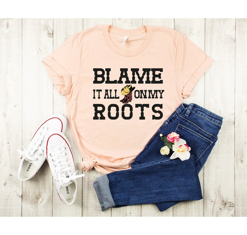 May include: A peach-colored t-shirt with the text "Blame it all on my roots" in black lettering. A cartoon boot with a cowboy hat is in the middle of the text. The shirt is laid out on a wooden surface with a pair of white sneakers and blue jeans.