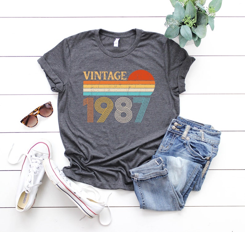 Peut inclure: Un t-shirt gris fonc&eacute; avec un design vintage 1987 repr&eacute;sentant un coucher de soleil avec des rayures orange, jaunes, bleues et vertes. Le t-shirt est pos&eacute; &agrave; plat sur une surface en bois blanc avec une paire de baskets blanches, un jean bleu et des lunettes de soleil.